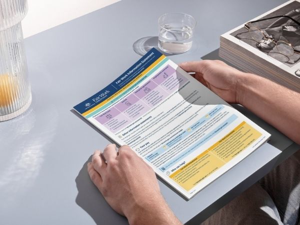 Hands holding a printed Fair Work Information Statement on a desk with a glass of water and reading glasses nearby.